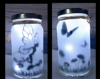 May include: A white frosted glass jar with a fairy silhouette and dandelion design. The jar is lit from within, creating a warm glow. The jar has a brown twine tied around the lid and the words "The Gift Page" are printed on the bottom.