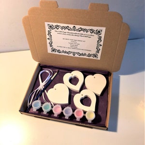 Op de afbeelding: Een knutselset voor het decoreren van keramische harten. De set bevat vier witte keramische harten met gaten om op te hangen, zes kleine verfpotjes, een kwast en een wit lint. De set is verpakt in een bruine kartonnen doos met een paarse tissuepapier voering.