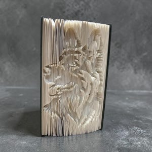 May include: A book with pages carved to create a wolf's head profile. The pages are light beige, and the edges are black. The wolf's features are detailed, with visible fur and facial structure. The book is standing upright.