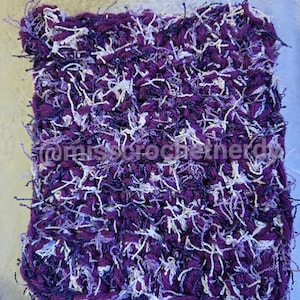 May include: A square, hand-crocheted scrubby featuring a textured, shaggy design in shades of deep purple, light purple, and white yarn. The text @misscrochetherdy is clearly visible across the center of the piece in a white, sans-serif font.