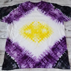 May include: A white t-shirt with a tie-dye design in shades of purple, yellow, and black. The design is a diamond shape with a yellow center and purple and black edges.