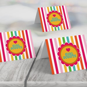 May include: Three colorful place cards with a striped background and a circle with a heart and the text "Girl Scouts".