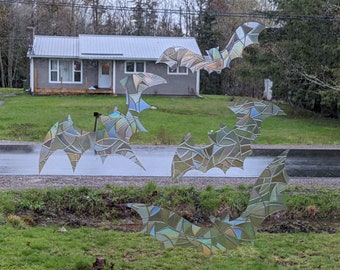 May include: Five iridescent bat window clings, each with a stained glass design, are displayed on a window. The bats are arranged in a cluster, with their wings spread out. The background is a blurry image of a house and trees.