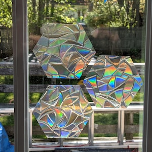 May include: Three iridescent hexagonal window clings with a geometric pattern. The clings are made of a clear material with a rainbow effect.