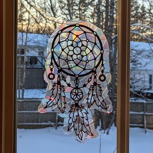 Könnte beinhalten: Ein schwarz-weißer Traumfänger mit schillernden Federn und Perlen. Der Traumfänger befindet sich an einem Fenster mit schneebedecktem Hintergrund.