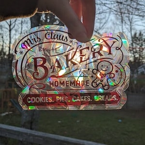May include: A holographic sticker with a red and white design. The sticker says "Mrs. Claus Bakery Est. 1845" and "Homemade Cookies. Pies. Cakes. Breads."
