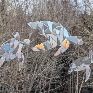 Könnte beinhalten: Drei Silhouetten von Schildkröten aus Buntglas in Grautönen, Blau und Orange vor einem Hintergrund aus braunen Zweigen.