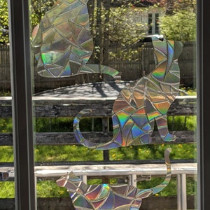 May include: Three iridescent cat silhouettes are displayed on a window. The cats are made of a mosaic of small, iridescent pieces. The cats are arranged in a staggered formation, with the largest cat at the top and the smallest cat at the bottom.