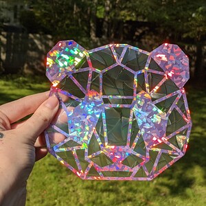 May include: A holographic iridescent bear head sticker with a geometric design. The sticker is made of a clear material with a holographic finish. The bear's head is facing forward and has a pink outline.