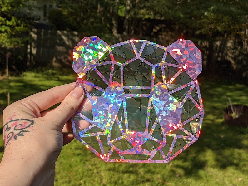 May include: A holographic iridescent bear head sticker with a geometric design. The sticker is made of a clear material with a holographic finish. The bear's head is facing forward and has a pink outline.