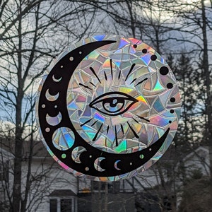 May include: A black and white crescent moon with a single eye inside. The moon is surrounded by a rainbow iridescent background.