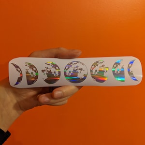 May include: A white sticker sheet with a holographic finish featuring a series of moon phases, including a full moon, half moon, crescent moon, and new moon.