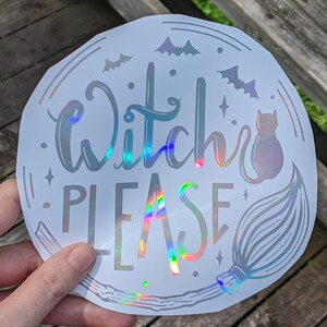 May include: A round, holographic sticker with the words "Witch PLEASE" in a whimsical font. The design includes bats, stars, a black cat, and a broom, all in a silver color. The sticker has a glossy finish and is held by a hand.