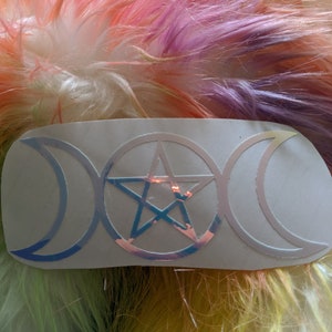 Puede incluir: Una pegatina plateada con un símbolo de triple luna y un pentagrama. La pegatina está sobre un fondo blanco y tiene un acabado holográfico.