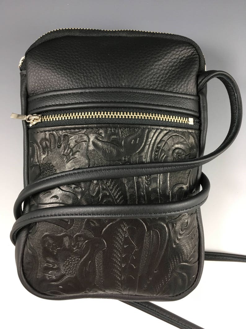 May include: Black leather crossbody bag with an embossed floral design. The bag has a zipped pocket on the front and a long adjustable strap.