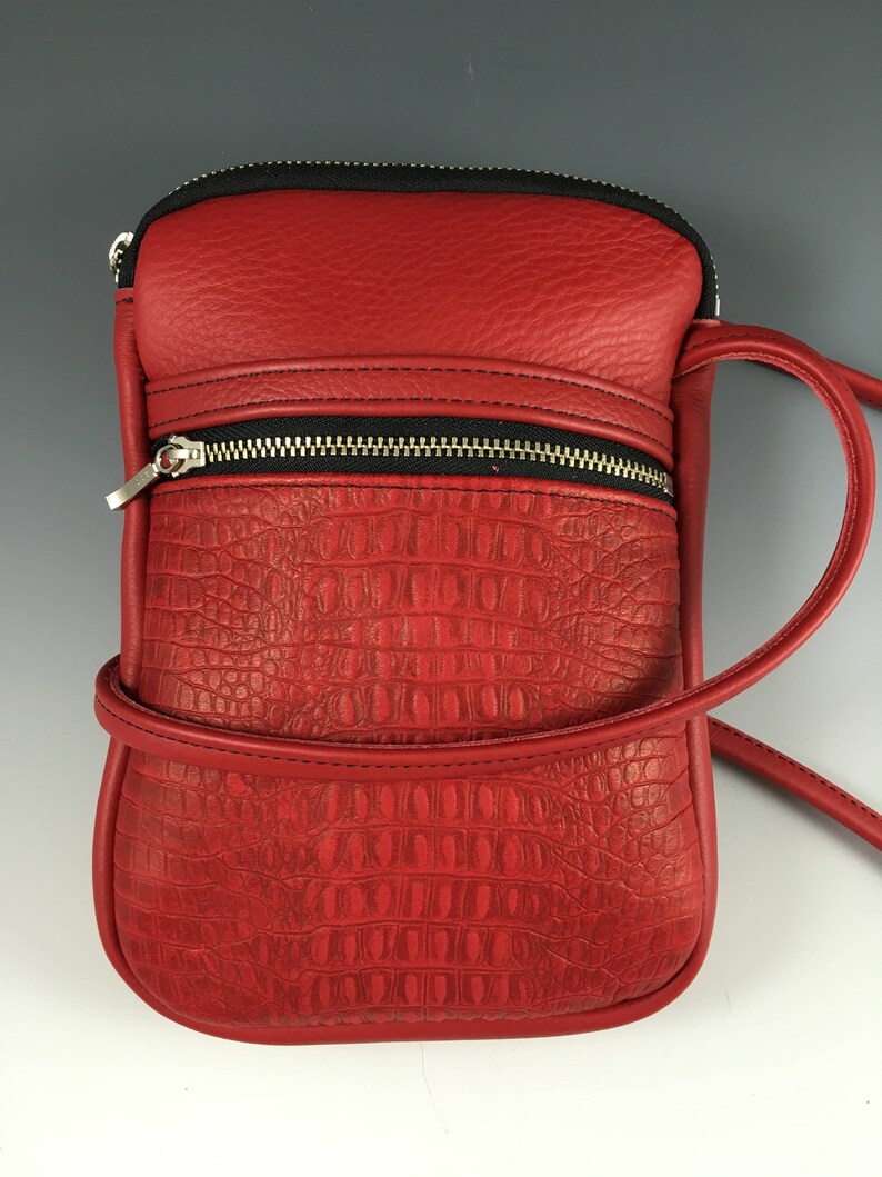 May include: A red leather crossbody bag with a crocodile embossed pattern and a black zipper. The bag has a long strap and a small pocket on the front.