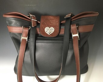 May include: A black leather tote bag with brown leather trim and straps. The bag has a heart-shaped metal accent on the front.