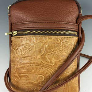 May include: A brown leather crossbody bag with a tooled floral design. The bag has a zipped pocket on the front and a brown adjustable strap.