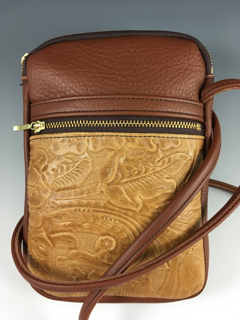 May include: A brown leather crossbody bag with a tooled floral design. The bag has a zipped pocket on the front and a brown adjustable strap.