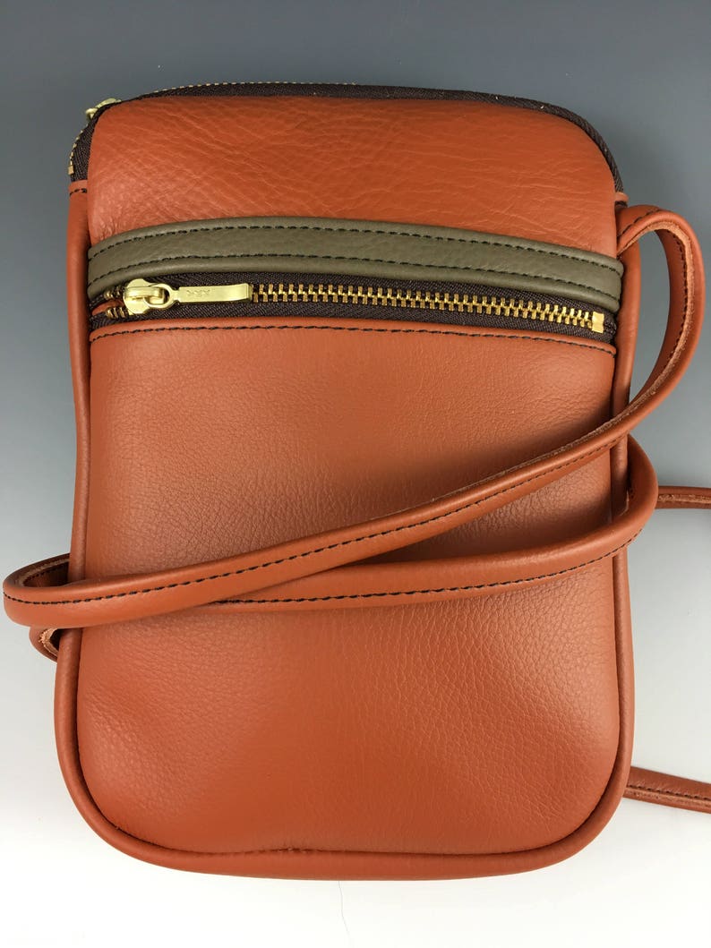 May include: A brown leather crossbody bag with a zipped pocket and a brown and grey trim. The bag has a long adjustable strap.
