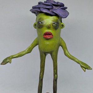 May include: A quirky sculpture of a humanoid figure. It has a green body, red lips, and a purple flower-like headpiece. The figure stands on a black base with arms outstretched. The sculpture is hand-painted.