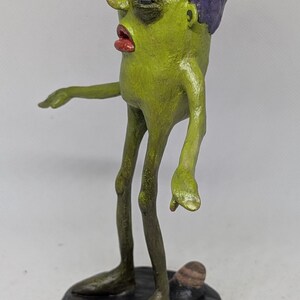 May include: A handmade sculpture of a green figure with a purple flower-like headpiece and red lips. The figure stands on a black base, with thin legs and arms outstretched. The sculpture has a unique, artistic style.