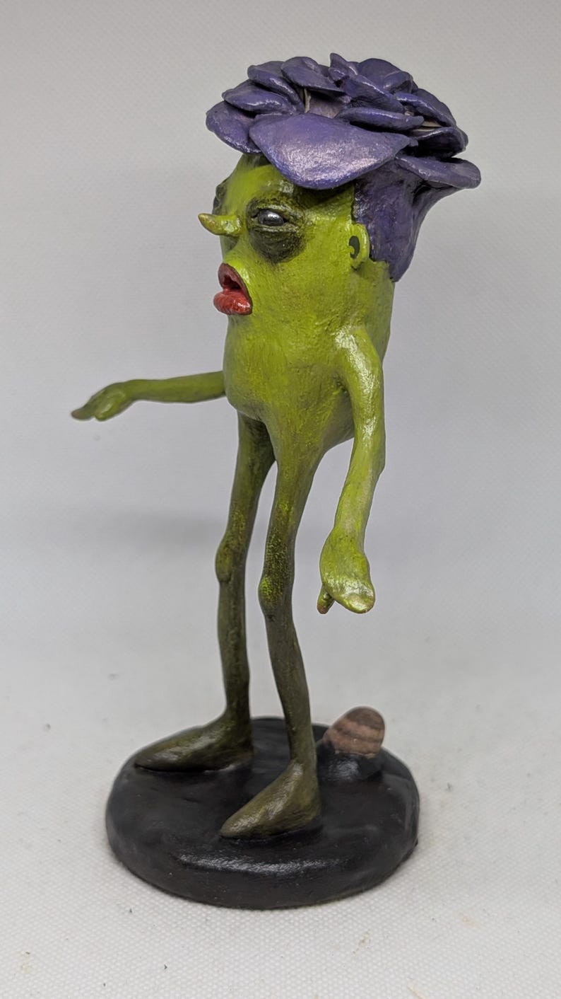 May include: A handmade sculpture of a green figure with a purple flower-like headpiece and red lips. The figure stands on a black base, with thin legs and arms outstretched. The sculpture has a unique, artistic style.