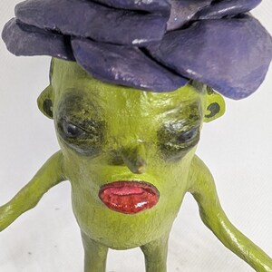 May include: A quirky, handmade figurine of a green creature with a purple flower-like headpiece and red lips. The figure has outstretched arms and stands on a black base, showcasing a unique, artistic design.