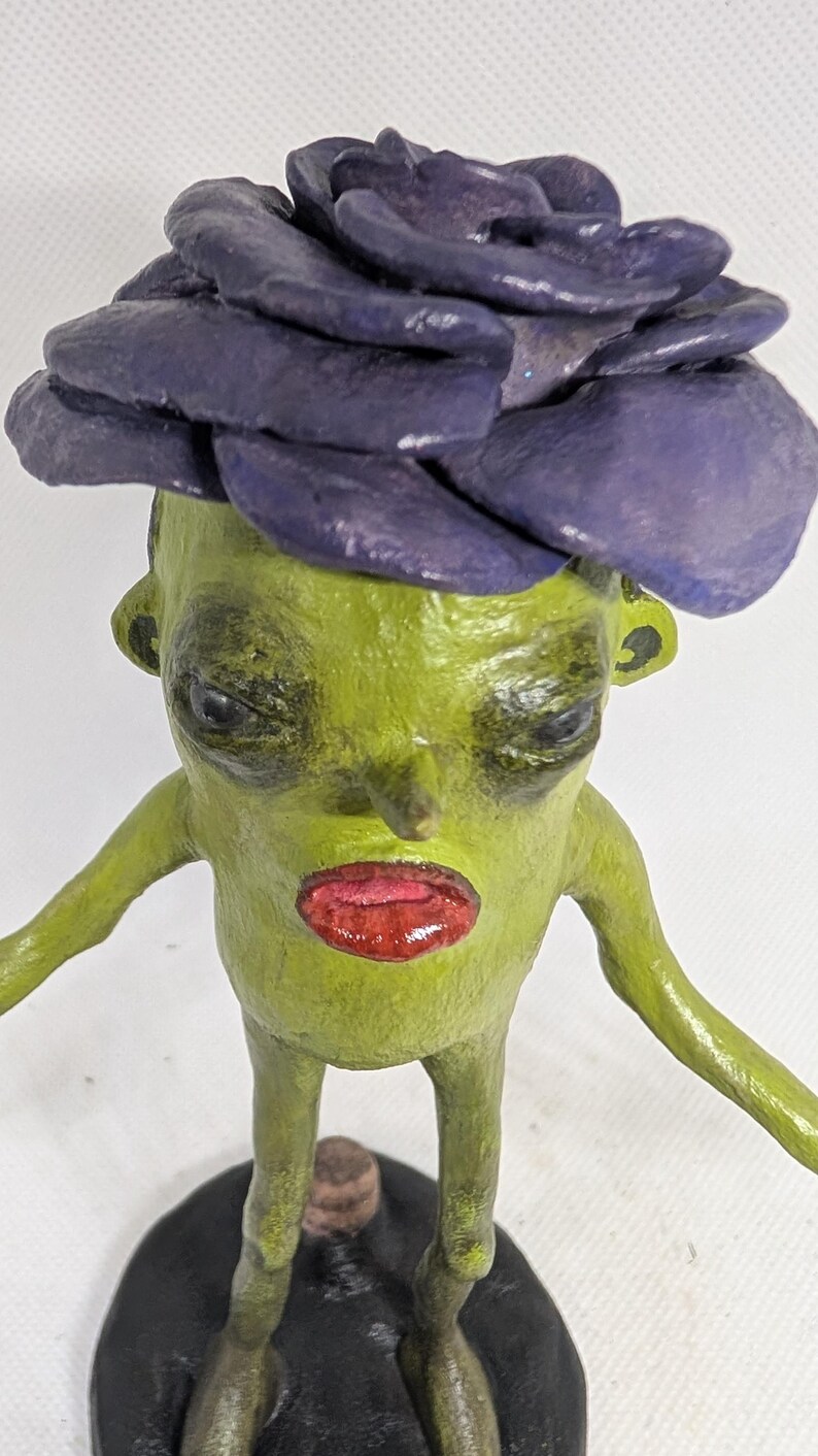 May include: A quirky, handmade figurine of a green creature with a purple flower-like headpiece and red lips. The figure has outstretched arms and stands on a black base, showcasing a unique, artistic design.