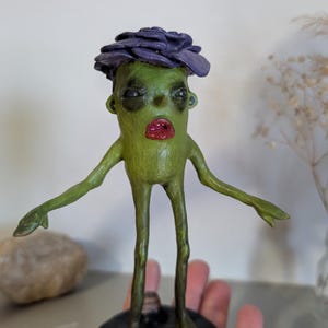 May include: A quirky green sculpture of a humanoid figure with a purple flower-like headpiece and red lips. The figure has outstretched arms and stands on a black circular base. The sculpture is held in a hand.
