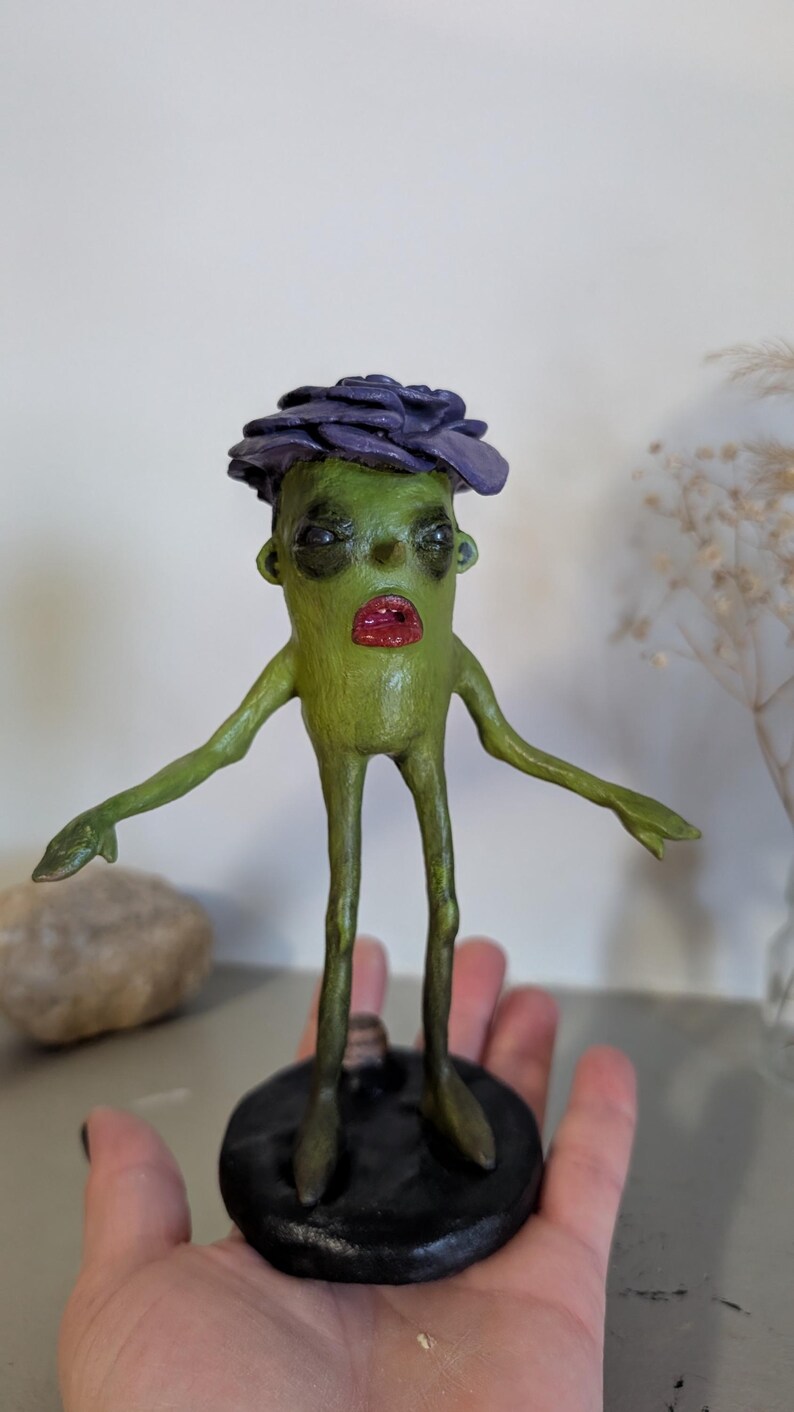 May include: A quirky green sculpture of a humanoid figure with a purple flower-like headpiece and red lips. The figure has outstretched arms and stands on a black circular base. The sculpture is held in a hand.