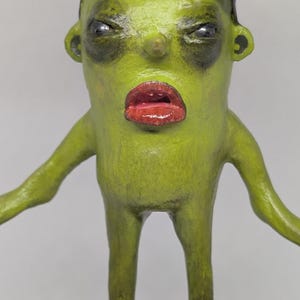May include: A quirky sculpture of a humanoid figure with a green complexion, red lips, and a purple flower-like headpiece. The figure has a stern expression and outstretched arms, with a detailed, textured finish.