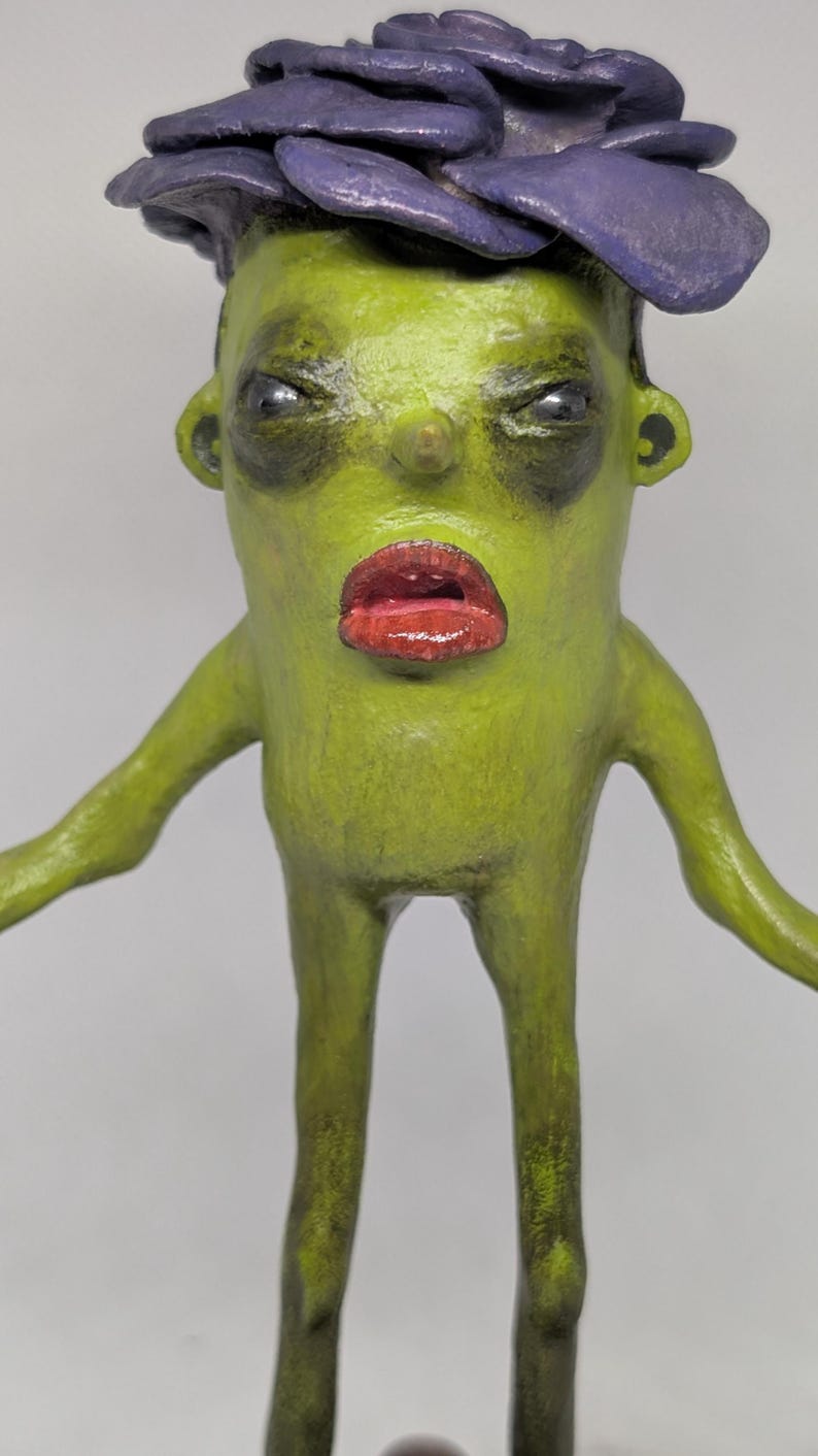 May include: A quirky sculpture of a humanoid figure with a green complexion, red lips, and a purple flower-like headpiece. The figure has a stern expression and outstretched arms, with a detailed, textured finish.