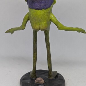 May include: A quirky, handmade sculpture of a creature with a green body, long legs, and outstretched arms. It has a purple flower-like head and stands on a dark, round base. The sculpture is a unique piece of art.