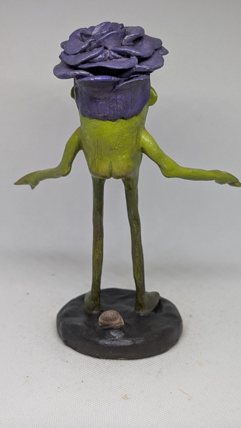 May include: A quirky, handmade sculpture of a creature with a green body, long legs, and outstretched arms. It has a purple flower-like head and stands on a dark, round base. The sculpture is a unique piece of art.