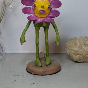 May include: A whimsical, handmade flower figurine with a purple and yellow face and petals. The figure has green arms and legs, standing on a brown circular base. The sculpture has a unique, expressive design.