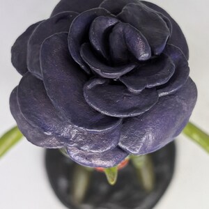 May include: A dark purple rose, viewed from above. The petals are layered and textured. The stem is green, and the base is black. The background is white, creating a contrast.