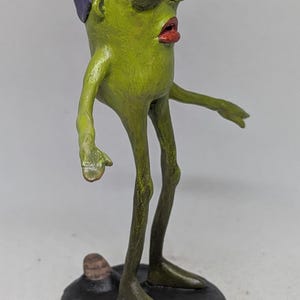 May include: A quirky, handmade sculpture of a humanoid figure. It has a green body, red lips, and a purple flower-like head. The figure stands on a black base, with thin legs and arms outstretched.