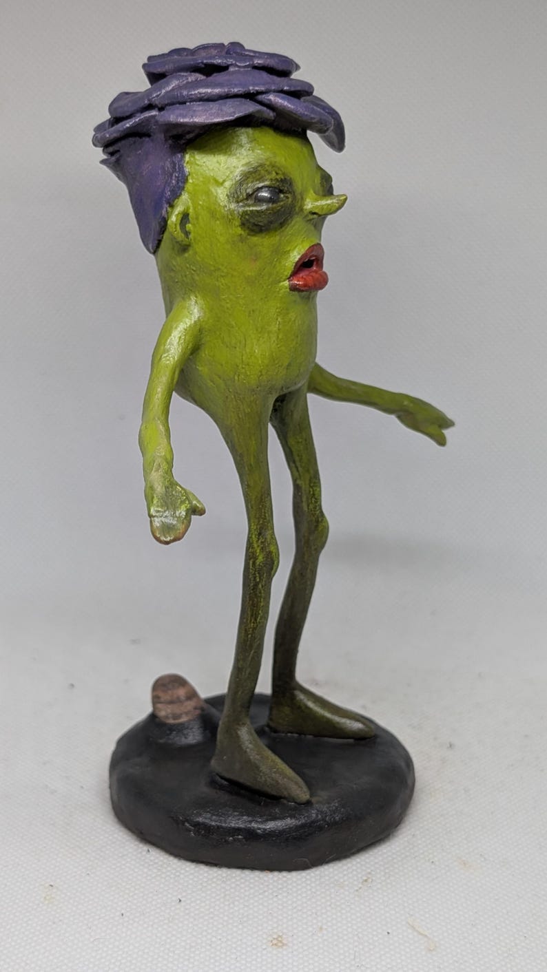 May include: A quirky, handmade sculpture of a humanoid figure. It has a green body, red lips, and a purple flower-like head. The figure stands on a black base, with thin legs and arms outstretched.