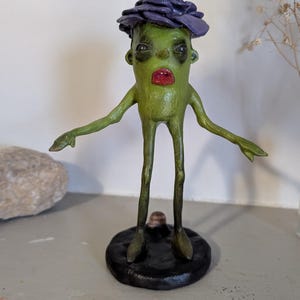 May include: A quirky, handmade sculpture of a green figure with a purple flower-like headpiece and red lips. The figure has outstretched arms and stands on a black circular base. The sculpture is a unique piece of art.