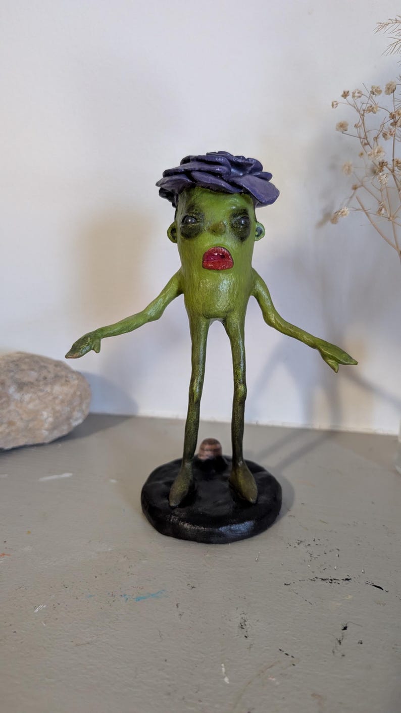 May include: A quirky, handmade sculpture of a green figure with a purple flower-like headpiece and red lips. The figure has outstretched arms and stands on a black circular base. The sculpture is a unique piece of art.