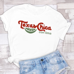 Puede incluir: Camiseta blanca con un gr&aacute;fico rojo y verde que dice "Texas Chica con lima". El gr&aacute;fico presenta una rodaja de lima.