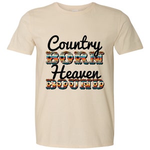 May include: A beige t-shirt with the text "Country Born Heaven Bound" in black lettering. The text is decorated with a colorful southwestern-style pattern.