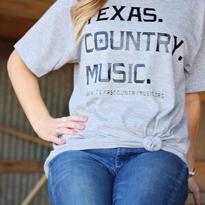 May include: A woman wearing a grey t-shirt with the text "TEXAS. COUNTRY. MUSIC." and a star graphic. The shirt also has the text "www.texascountrymusic.org". She is wearing blue jeans.