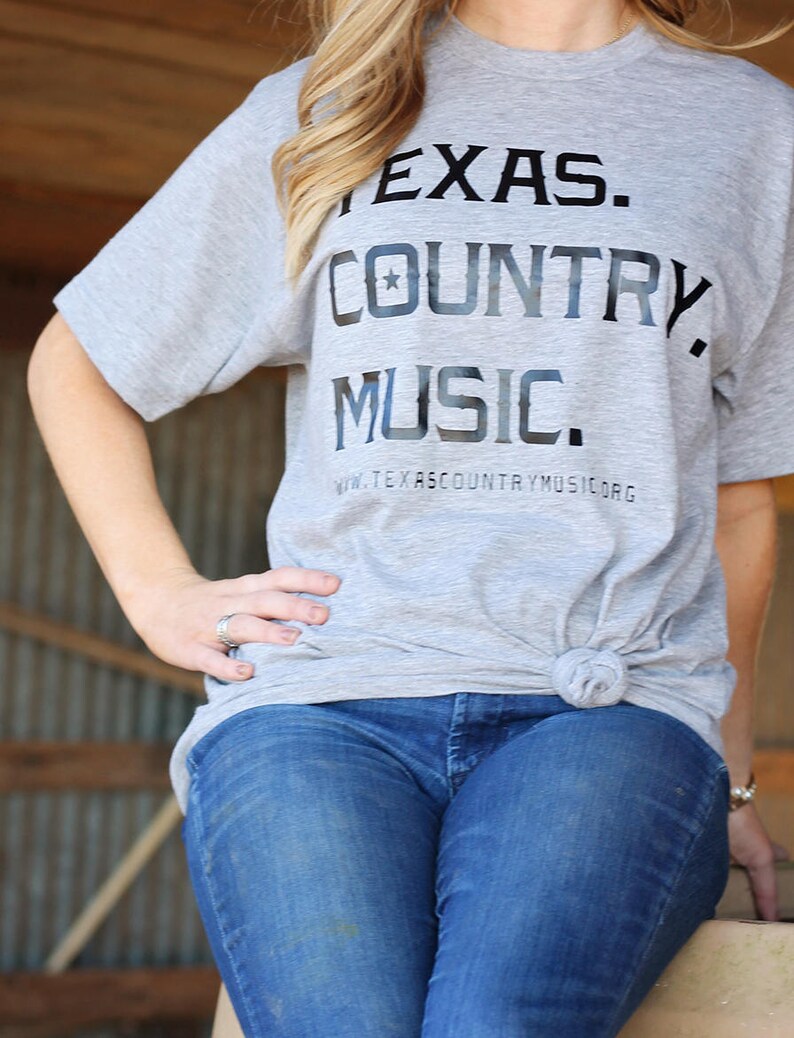 May include: A woman wearing a grey t-shirt with the text "TEXAS. COUNTRY. MUSIC." and a star graphic. The shirt also has the text "www.texascountrymusic.org". She is wearing blue jeans.