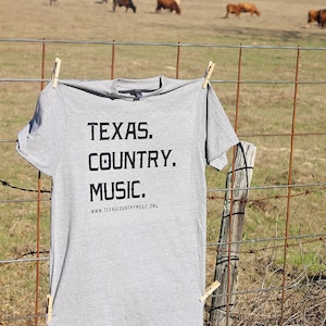 May include: A gray t-shirt with the text "TEXAS. COUNTRY. MUSIC." and the website "www.texascountrymusic.org" printed on it. The shirt is hanging on a rusty wire fence with wooden clothespins.