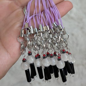 Pode incluir: Uma coleção de amuletos de contas com fechos prateados e cordões lavanda. Cada amuleto apresenta uma conta redonda transparente, uma conta cilíndrica preta e uma pequena conta vermelha. Os amuletos são projetados para uso como acessórios.