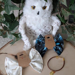 May include: A plush white owl with yellow eyes sits above two fabric hair bows and a brown braided bracelet with a gold charm. One bow is white with a gold wing pattern, the other is black and blue.
