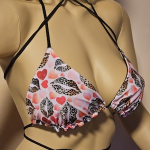 Op de afbeelding: Een halter bikinitop met een print van rode harten en luipaardlippen op een lichtroze achtergrond. De bikinitop heeft zwarte bandjes die om de nek en op de rug worden gestrikt.