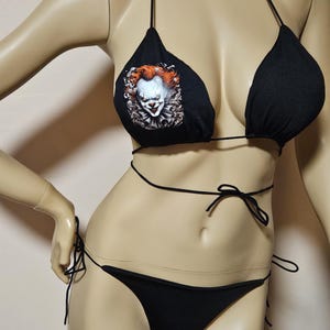 May include: Black bikini set featuring a string top and bottom. The top has a graphic of a scary clown with red hair and white face paint. The bikini is displayed on a mannequin.